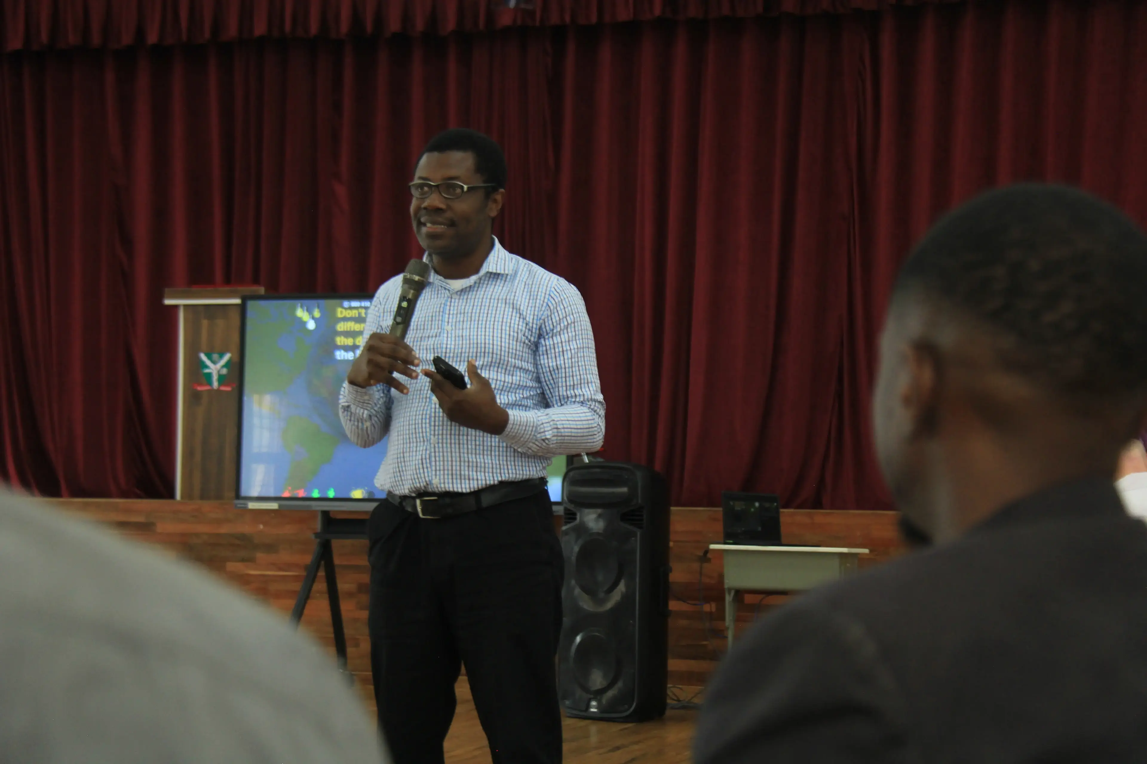Semiu Ajala presenting on stage to a room of teacher trainees, with a screen displaying content behind him